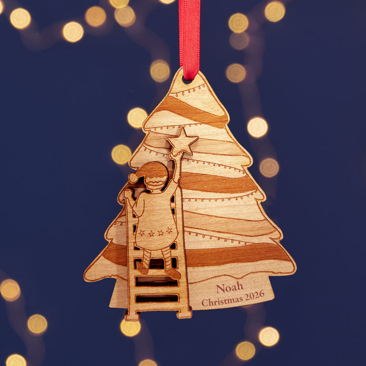 Personalised wooden Christmas tree decoration with a child climbing a ladder to put the star on the of the tree. The text reads Noah Christmas 2026. The background is dark blue with fairy lights.