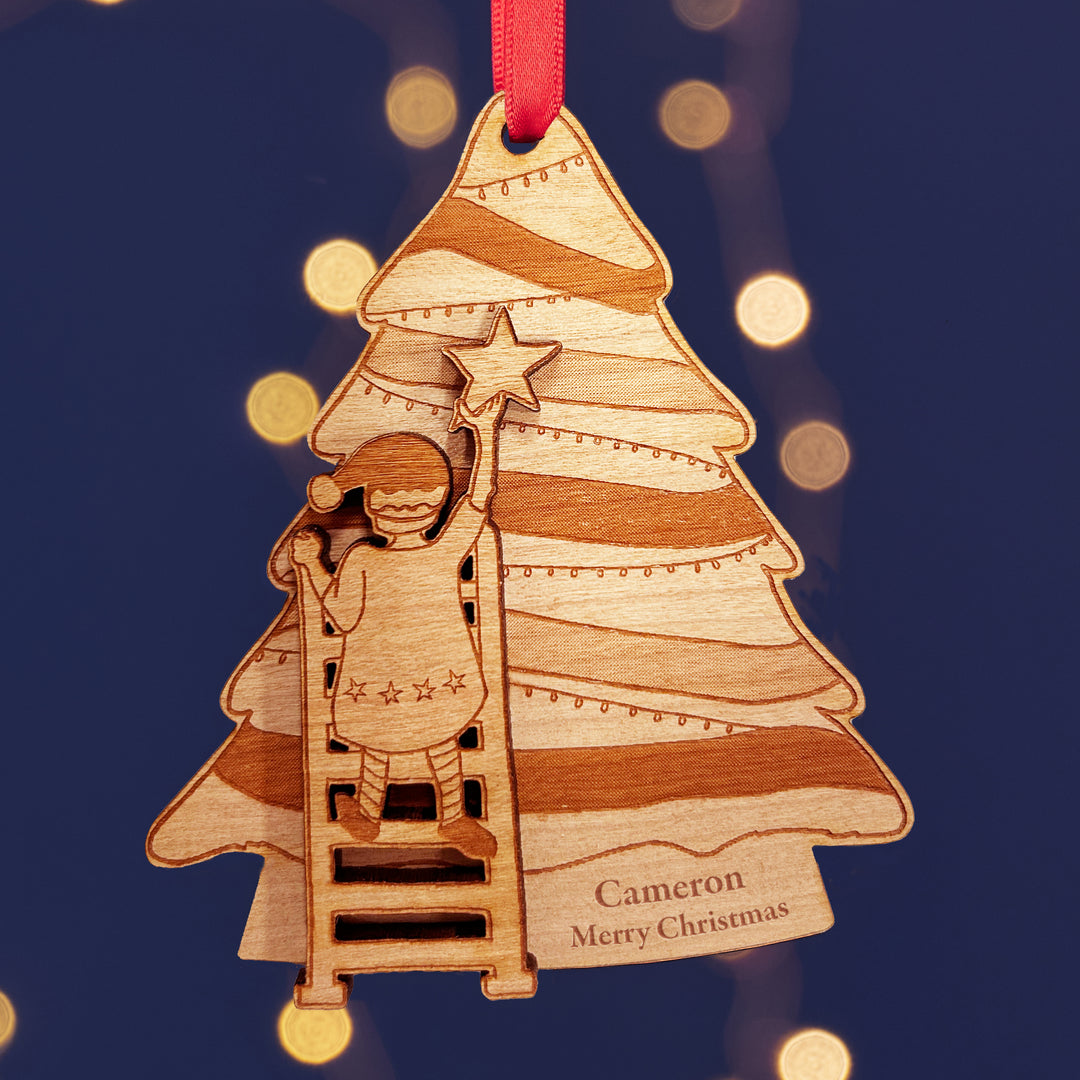 Personalised wooden Christmas tree decoration with a child climbing a ladder to put the star on the of the tree. The text reads Cameron Merry Christmas. The background is dark blue with fairy lights.