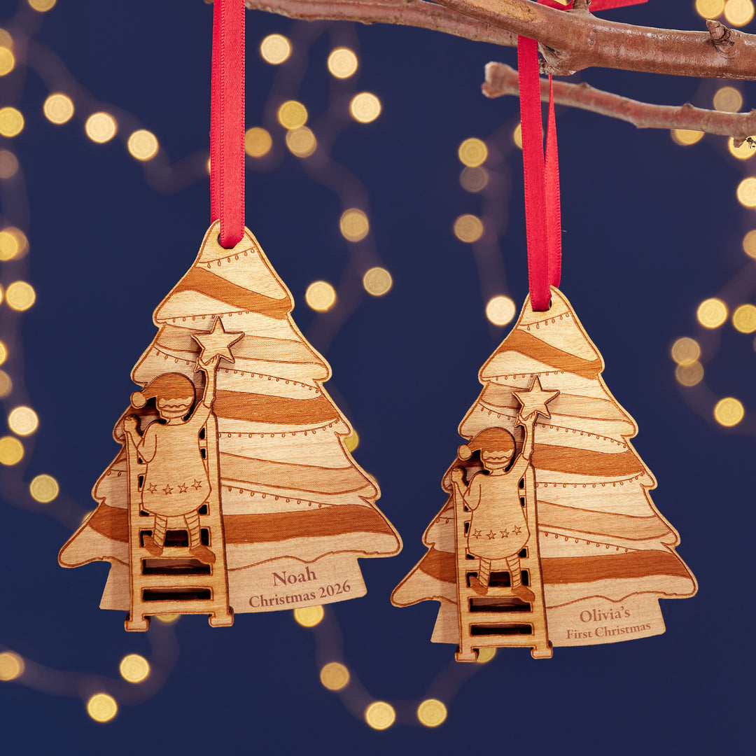  Two Personalised wooden Christmas tree decoration with a children climbing a ladder to put the star on the of the tree. The text on the left hand decoration reads Noah Christmas 2026 and the right hand decoration reads Olivia's first Christmas. The background is dark blue with fairy lights.