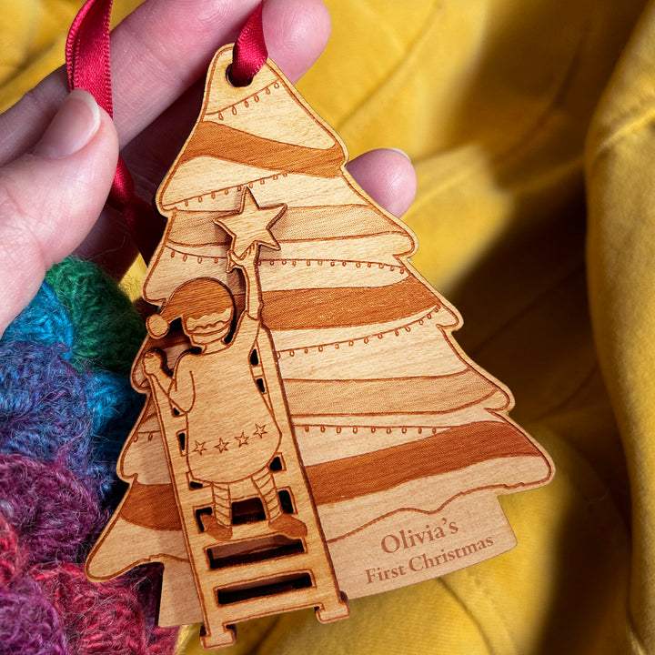 Wooden Christmas tree ornament with a child climbing a tree to hang a star, held by a hand against a yellow background.