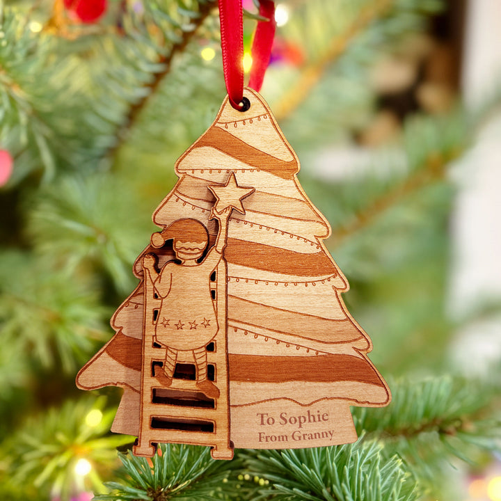 Wooden Christmas tree decoration shaped like a tree, with a child reaching for a star. The ornament has the text "To Sophie from Granny" and hangs from a red ribbon. The tree is decorated with lights.