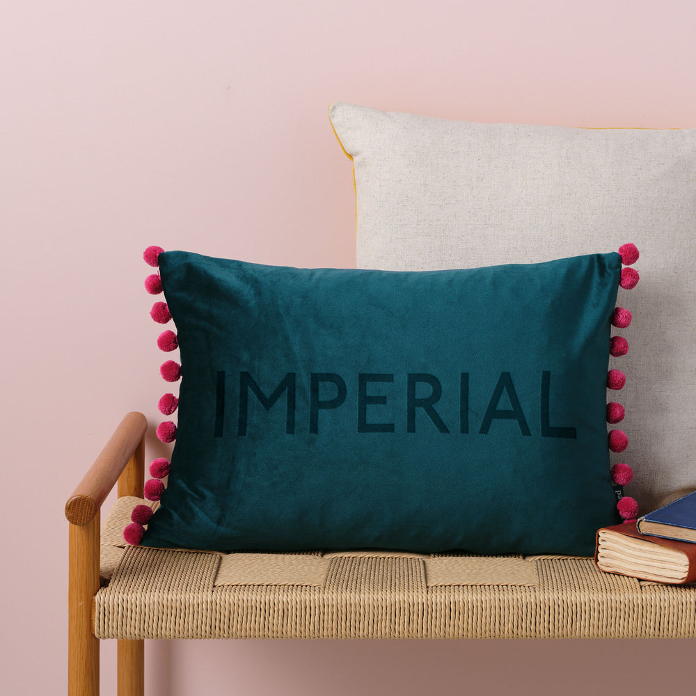 Imperial university graduation student gift, a teal pom pom cushion on a wicker bench against a pink wall.