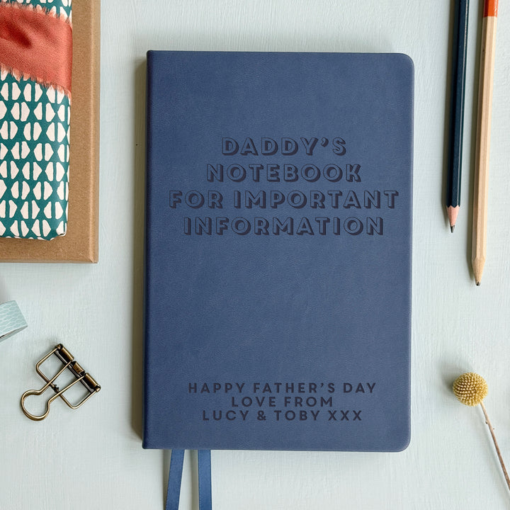 A top down image of a mid blue vegan leather notebook resting on a pale aqua table. The front of the notebook is engraved with the personalised text "DADDY'S NOTEBOOK FOR IMPORTANT INFORMATION" in a large font. HAPPY FATHER'S DAY LOVE FROM LUCY & TOBYXXX  is engraved in a smaller text at the bottom of the notebook. Surrounding the book are various stationary items and dried flowers.