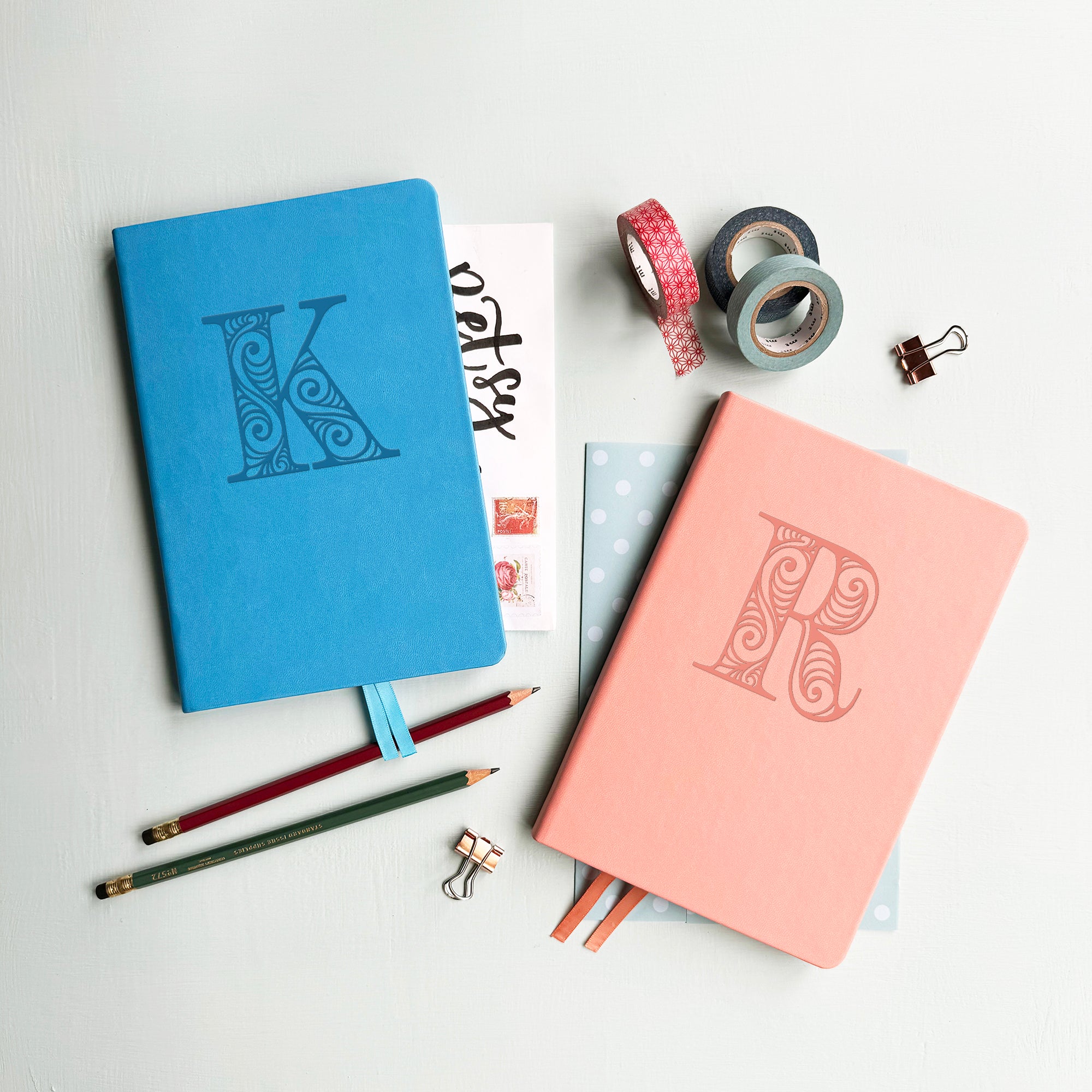 A top down image of two vegan leather notebooks, one blue and one pink. They are resting on a pale aqua table. The blue notebook is personalised with the letter K engraved in a large decorative font and the pink journal has the letter R.  Surrounding the book are various stationary items.
