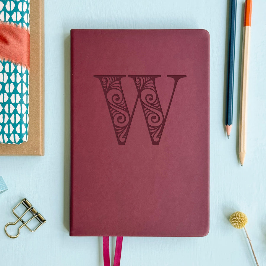 A top down image of a burgundy vegan leather notebook resting on a pale aqua table. The front of the notebook is personalised with the letter W engraved in a large decorative font. Surrounding the book are various stationary items.