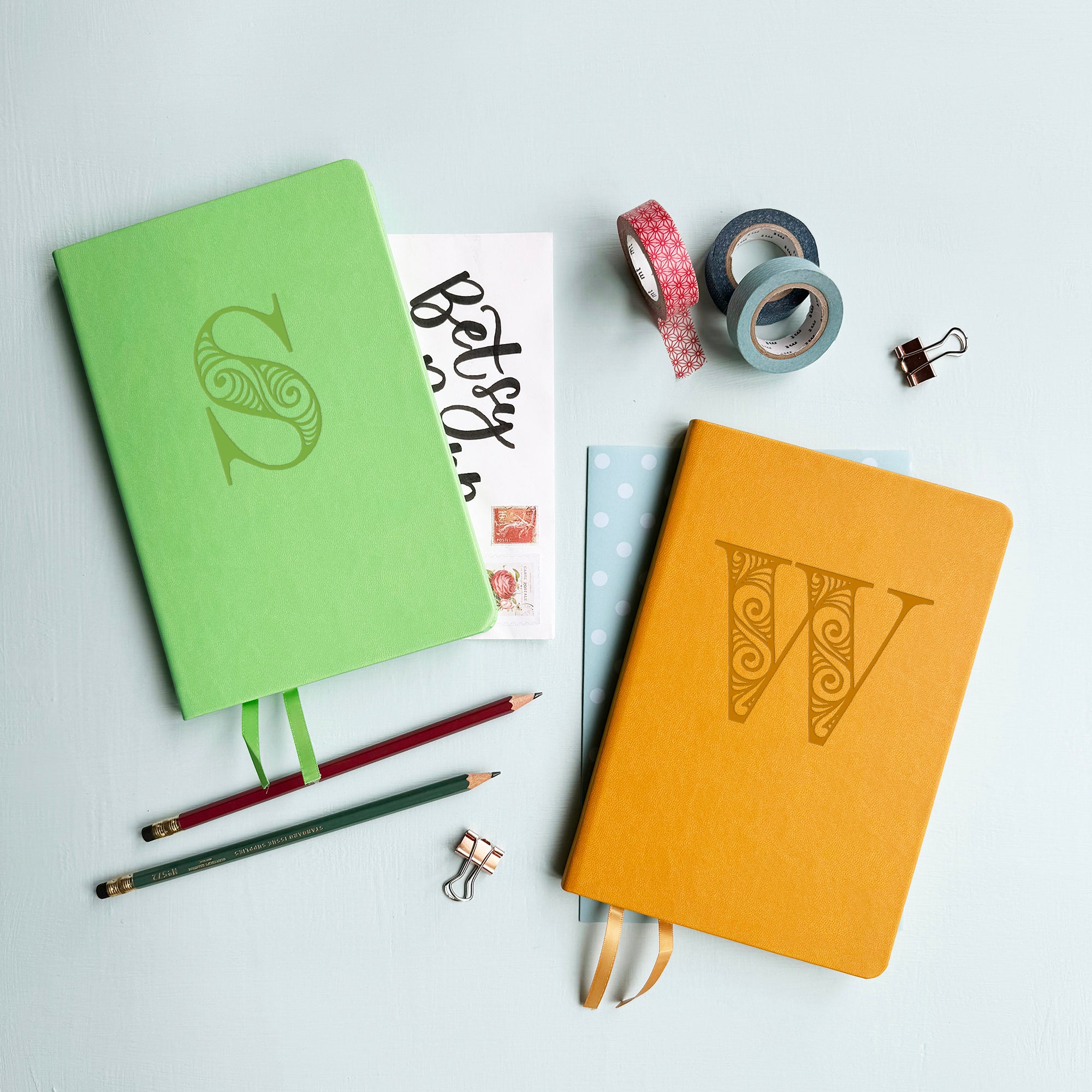 A top down image of two vegan leather notebooks, one green and one mustard yellow. They are resting on a pale aqua table. The green notebook is personalised with the letter S engraved in a large decorative font and the yellow journal has the letter W.  Surrounding the book are various stationary items.
