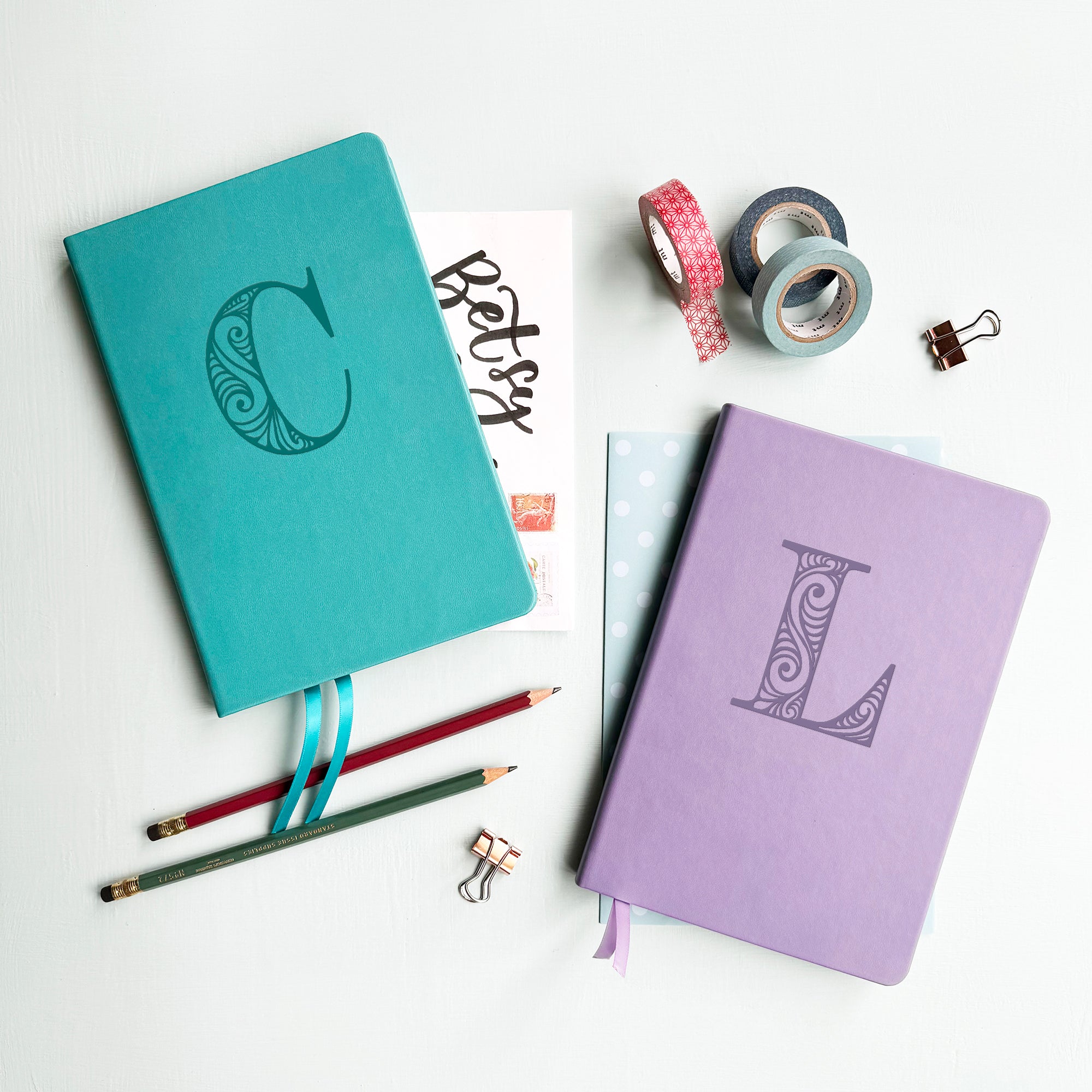 A top down image of two vegan leather notebooks, one turquoise and one lilac. They are resting on a pale aqua table. The turquoise notebook is personalised with the letter C engraved in a large decorative font and the lilac journal has the letter L.  Surrounding the book are various stationary items.
