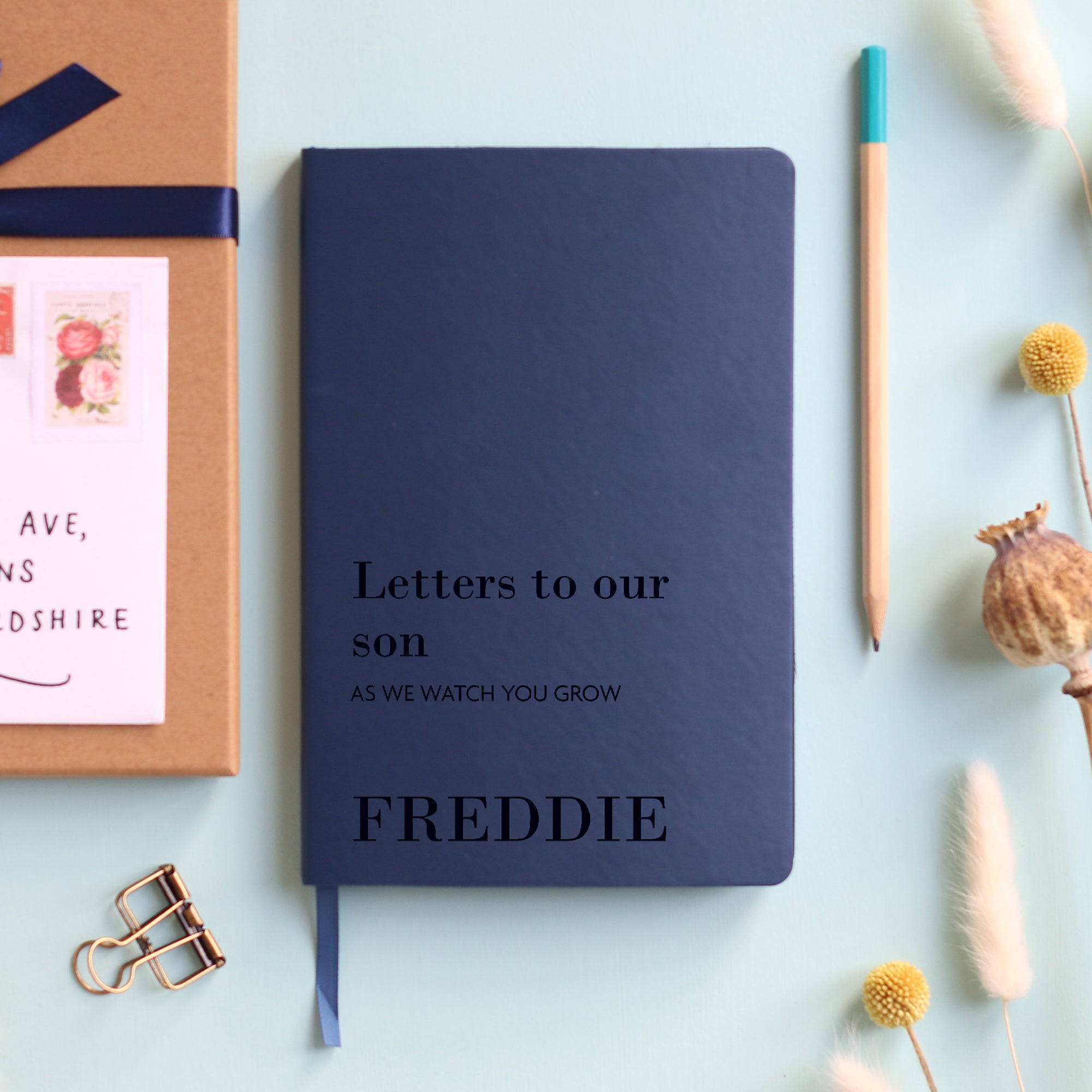 A top down image of a navy blue vegan leather notebook resting on a pale aqua table. The front of the notebook is engraved with the phrase Letters to our son in large text and underneath it is personalised with the words AS WE WATCH YOU GROW in a smaller font size. Personalised with the name Freddie. Surrounding the book are various stationary items.