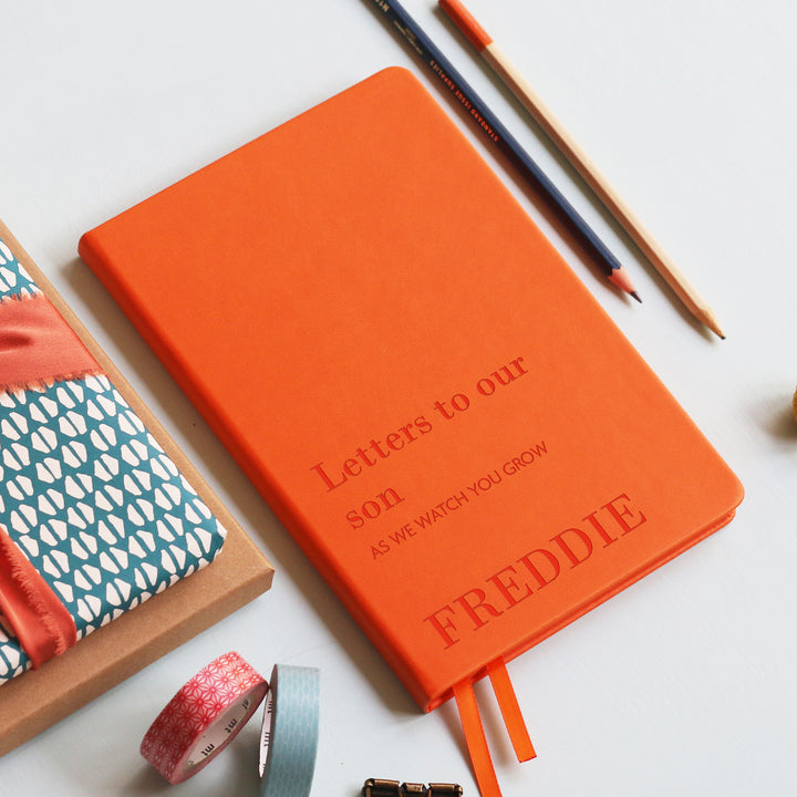 A top down image of a orange vegan leather notebook resting on a pale aqua table. The front of the notebook is engraved with the phrase Letters to our son in large text and underneath it is personalised with the words AS WE WATCH YOU GROW in a smaller font size. Personalised with the name Freddie. Surrounding the book are various stationary items.