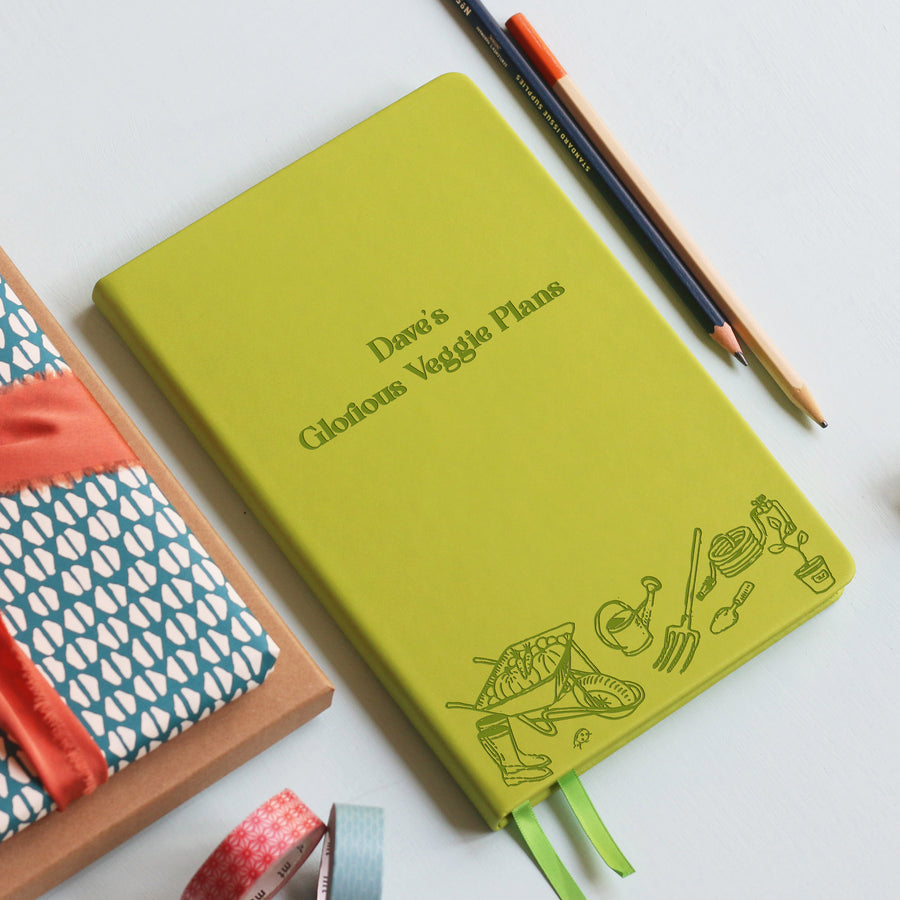 A top down image of a lime green vegan leather notebook resting on a pale aqua table. The front of the notebook features an engraving of gardening tools along the bottom edge. The personalised text reads "Dave's Glorious Veggie Plans". Surrounding the book are various stationary items and dried flowers.