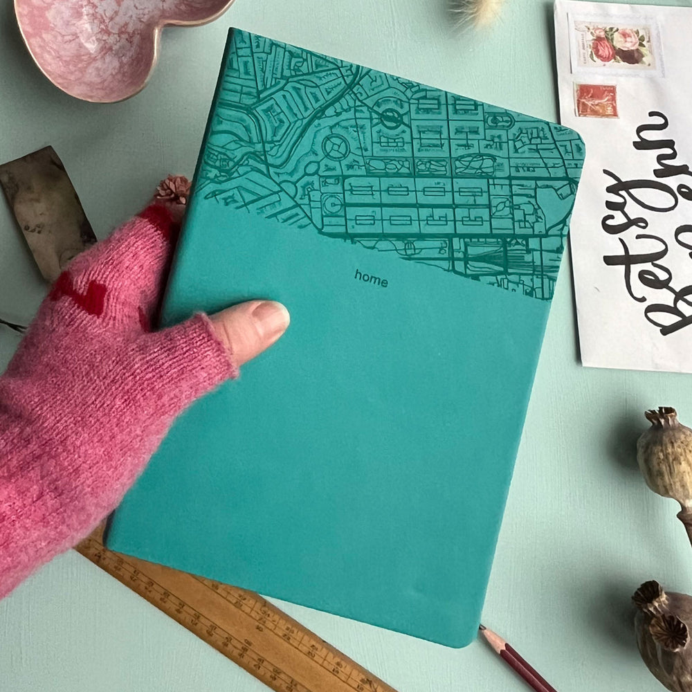 A close up image of a turquoise vegan leather notebook being held by a hand wearing a pink fingerless glove. The background is a pale aqua table. The top third of the notebook is engraved with a map and personalised with the text “home”. Surrounding the book are various stationary items and dried flowers.
