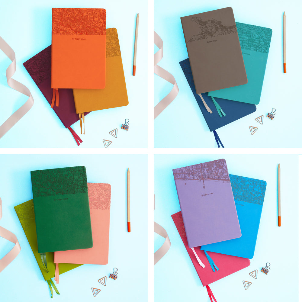 The image shows examples of personalised engraved text on all 12 colours of notebook. The books are stacked in in threes and are surround by various pieces of stationery