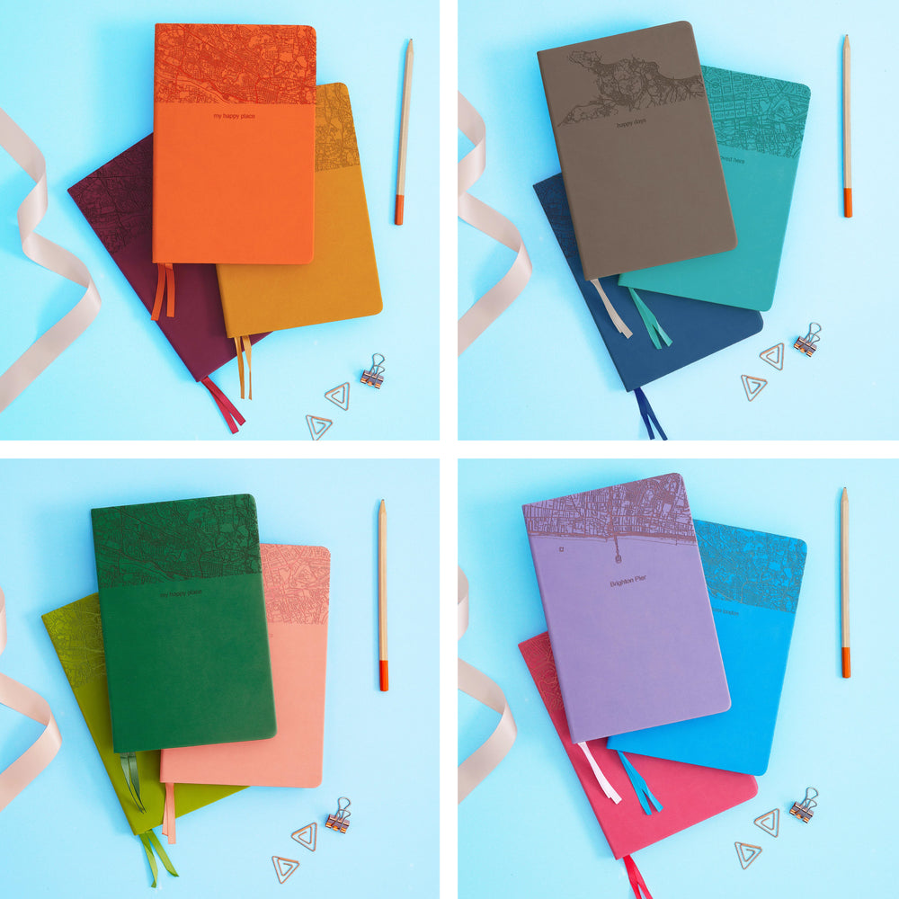 The image shows examples of personalised engraved text on all 12 colours of notebook. The books are stacked in threes and are surround by various pieces of stationery.
