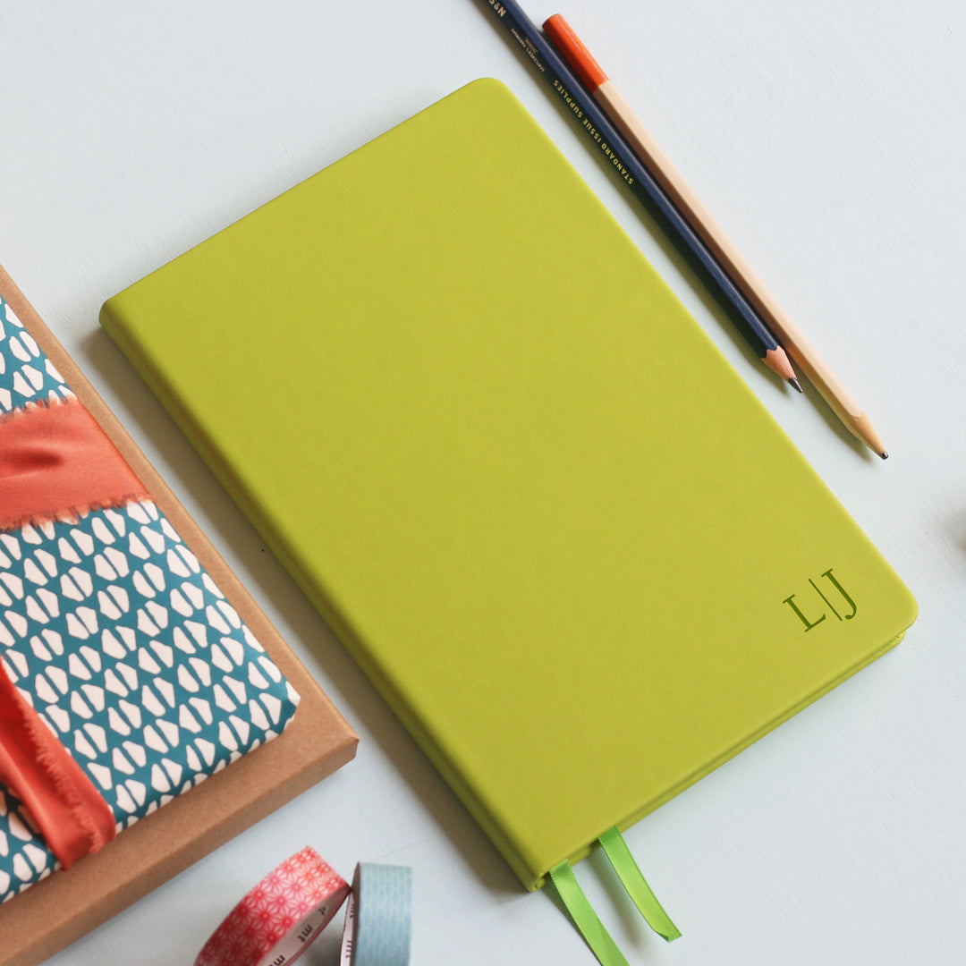 A5 lined lime green notebook lying on a pale blue background. Engraved on the notebook are the initials L|J in the bottom right corner. The background contains stationery and dried flowers.