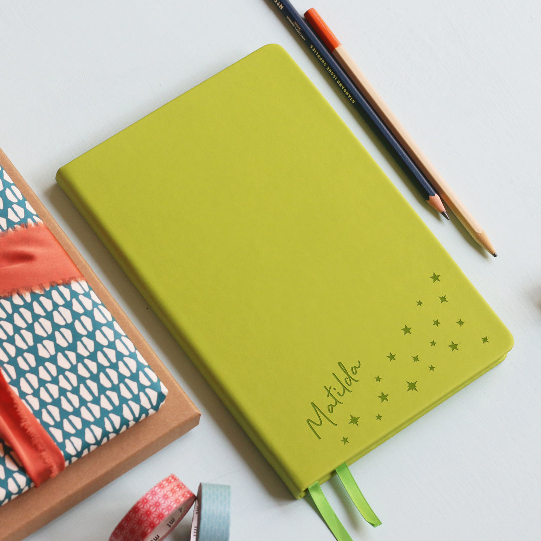 A flat lay image of a lime green vegan leather notebook resting on a pale aqua table. The front of the notebook is personalised with the name Matilda above a cluster of different sized engraved stars. Surrounding the book are various stationary items.