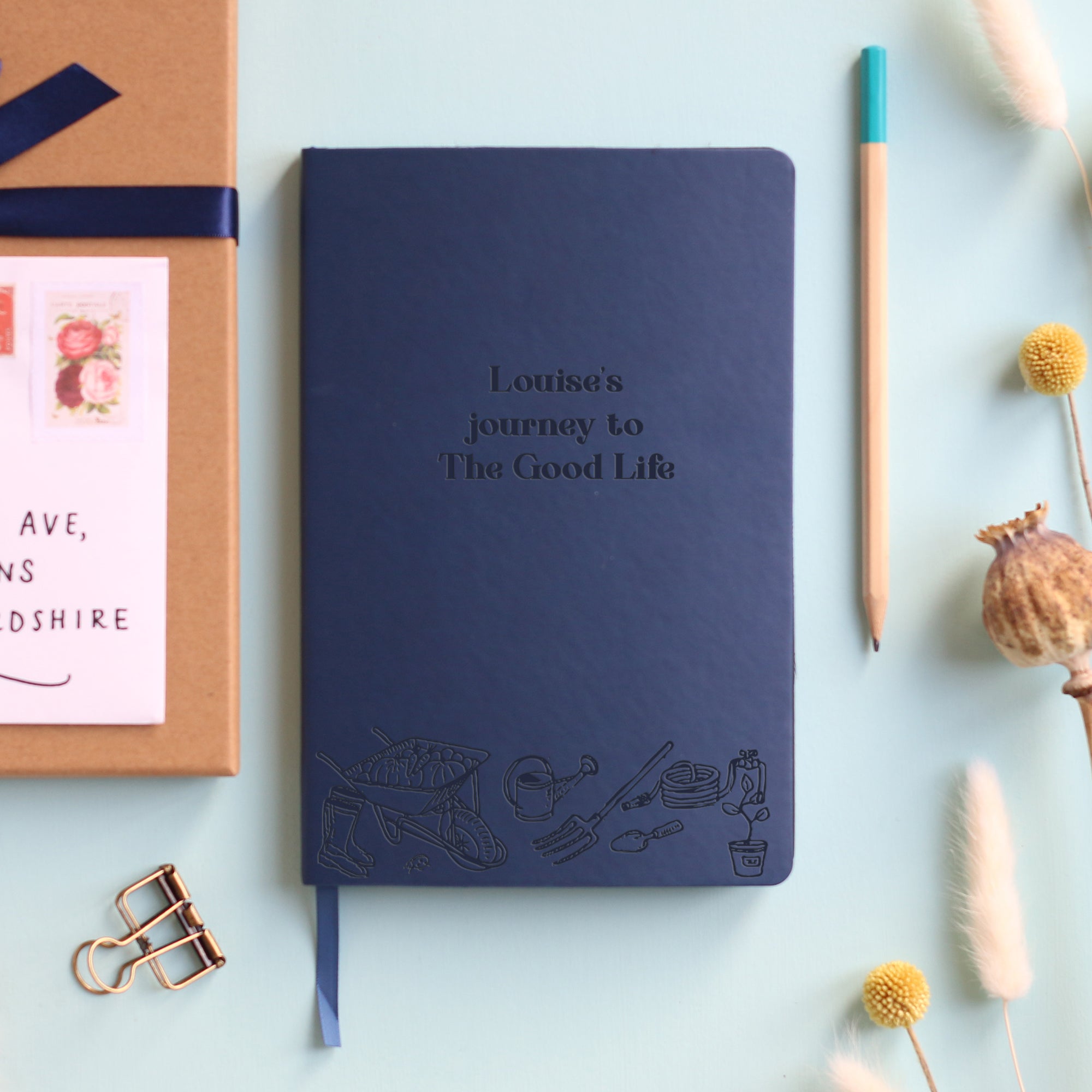 A top down image of a blue vegan leather notebook resting on a pale aqua table. The front of the notebook features an engraving of gardening tools along the bottom edge. The personalised text reads "Louise's journey to The Good Life". Surrounding the book are various stationary items and dried flowers.