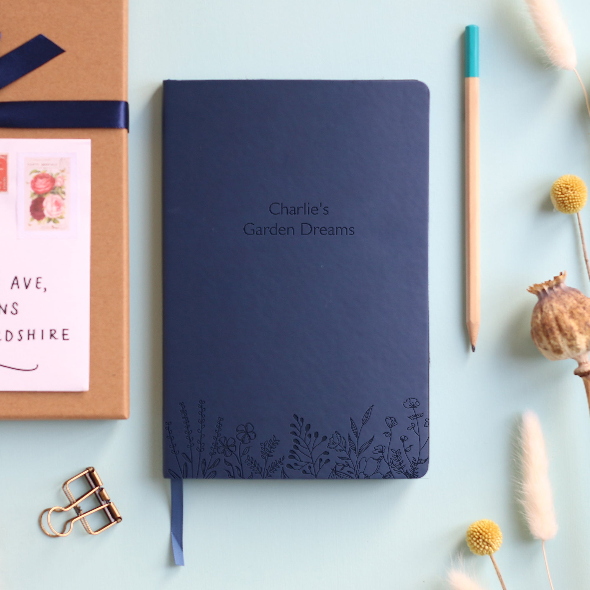 A top down image of a red vegan leather notebook resting on a pale aqua table. The front of the notebook features a pattern of delicate flowers along the bottom edge. The personalised engraving reads "Charlie's Garden Dreams". Surrounding the book are various stationary items and dried flowers.