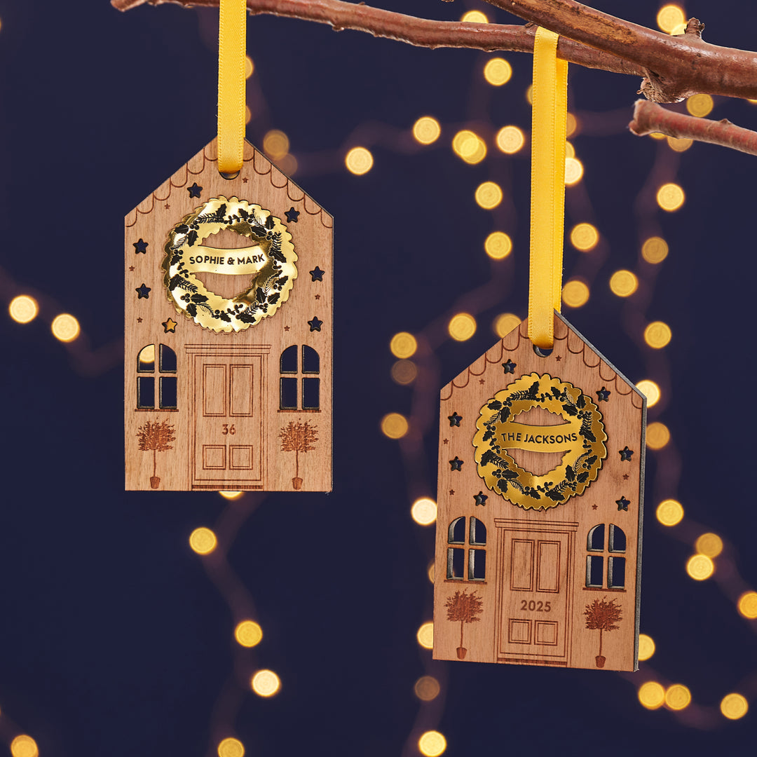 Wooden houses Christmas ornaments with a personalised golden wreath and engraved details hanging with a gold ribbon from a tree