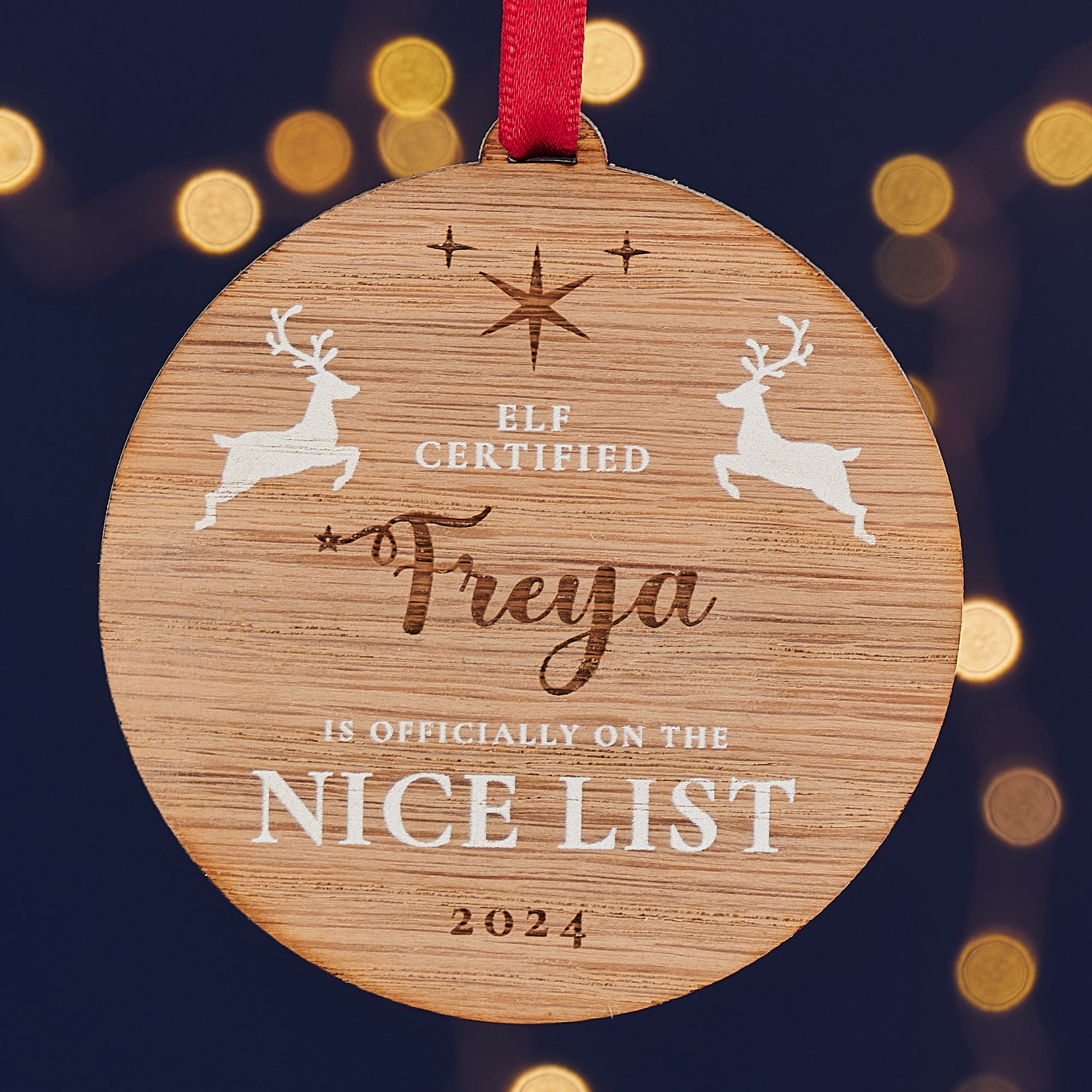 A round wooden Christmas ornament with the name "
Freya" engraved on it, along with the message "Elf Certified - Is Officially on the Nice List 2025" and two leaping reindeer, hanging on a Christmas tree.
