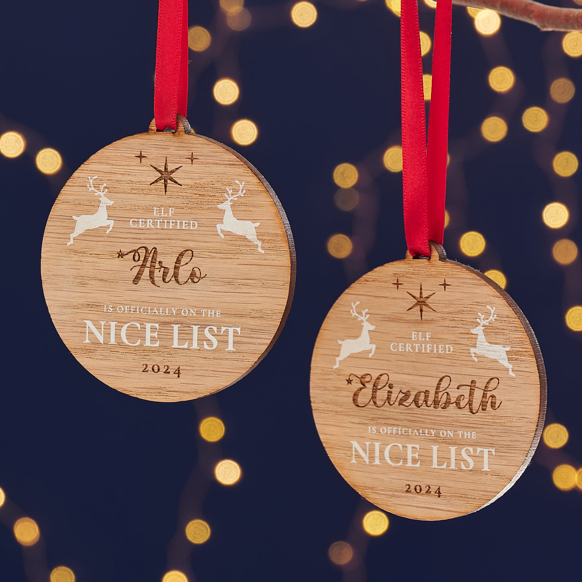 Two round wooden Christmas ornaments with the names "Arlo" and "Elizabeth" engraved on them, along with the message "Elf Certified - Is Officially on the Nice List 2024" and two leaping reindeer, hanging on a Christmas tree.