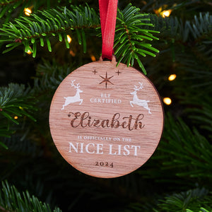 A round wooden Christmas ornament with the name "Elizabeth" engraved on it, along with the message "Elf Certified - Is Officially on the Nice List 2024" and two leaping reindeer, hanging on a Christmas tree.
