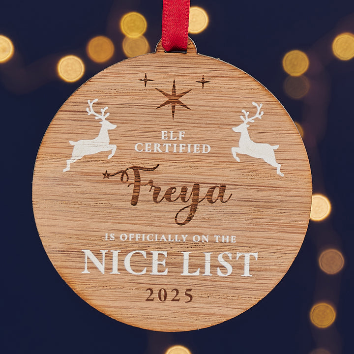 A round wooden Christmas ornament with the name "Freya" engraved on it, along with the message "Elf Certified - Is Officially on the Nice List 2025" and two leaping reindeer.
