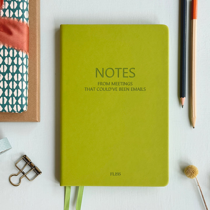 A top down image of a lime green vegan leather notebook resting on a pale aqua table. The front is engraved with the phrase "Notes from meetings that could’ve been emails" and personalised with the name Fliss. Surrounding the book are various.