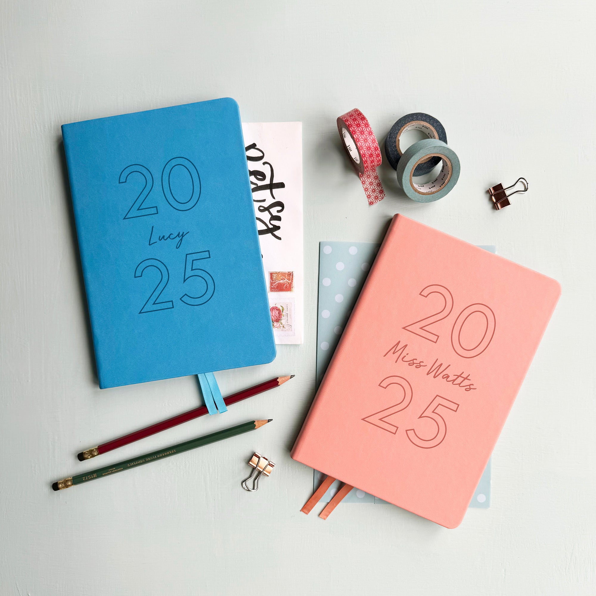 A top down image of two vegan leather notebooks, one blue and one pink. They are resting on a pale aqua table. Both are engraved with 2025. The blue notebook is personalised the name Lucy and the pink book features the name Miss Watts.  Surrounding the book are various stationary items.