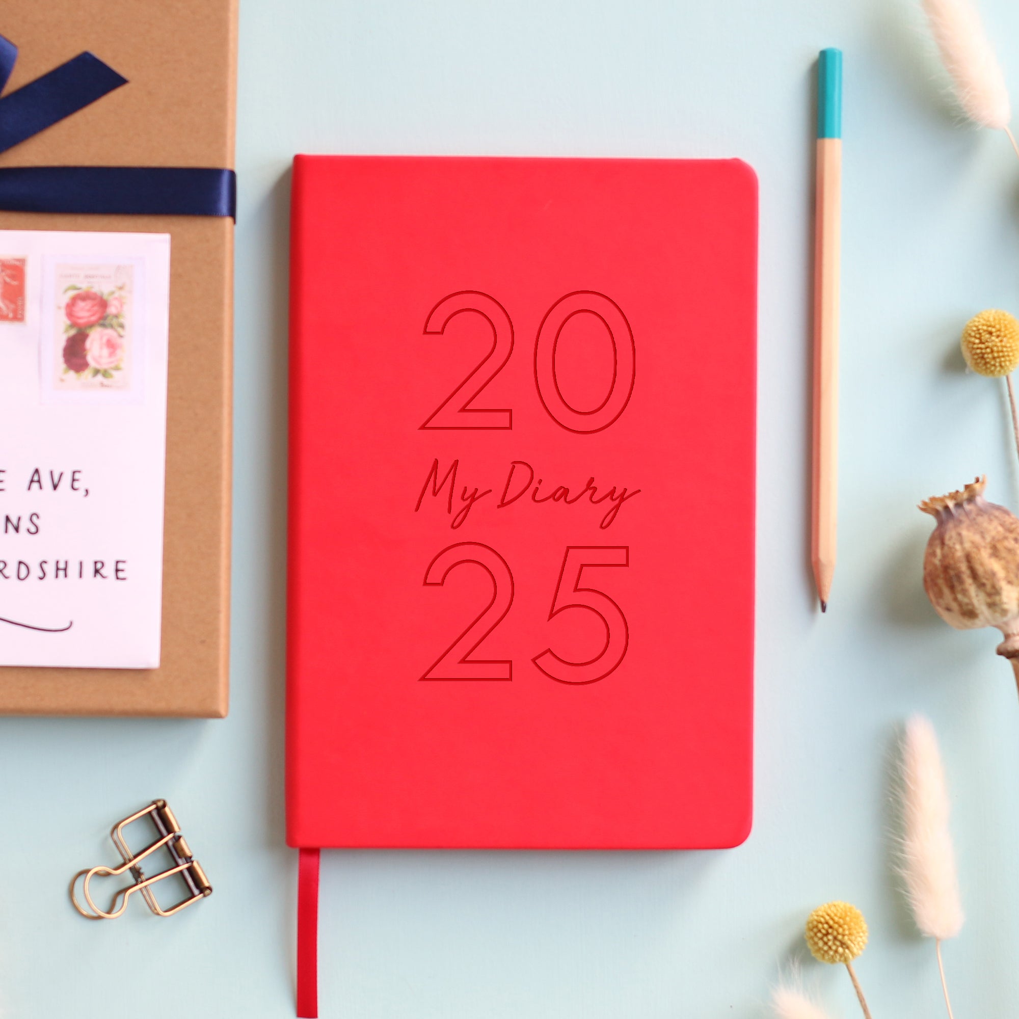 A top down image of a red vegan leather notebook resting on a pale aqua table. The front of the notebook is engraved with 2025 and the personalised text My Diary. Surrounding the book are various stationary items and dried flowers.