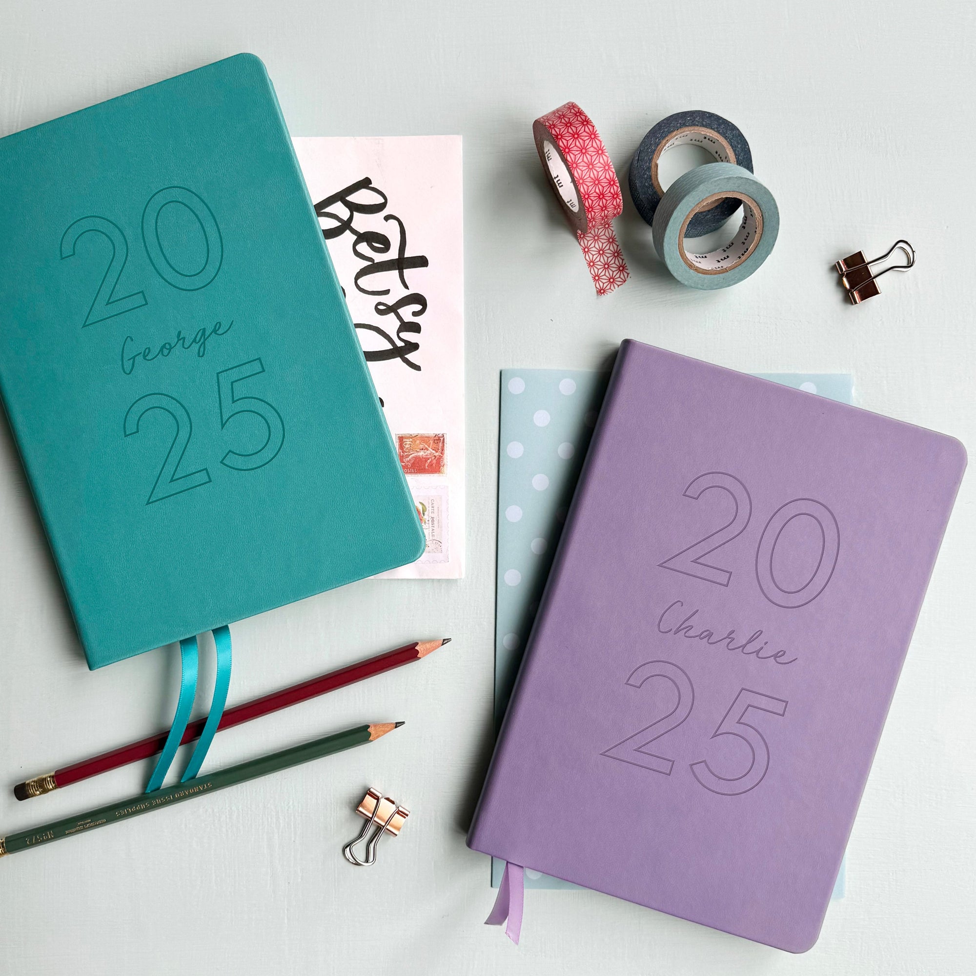 A top down image of a red vegan leather notebook resting on a pale aqua table. The front of the notebook is engraved with 2025 and the personalised text My Diary. Surrounding the book are various stationary items and dried flowers.