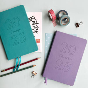 A top down image of two vegan leather notebooks, one turquoise and one lilac. They are resting on a pale aqua table. Both are engraved with 2025. The lilac notebook is personalised the name Charlie and the Turquoise book features the name George.  Surrounding the book are various stationary items.