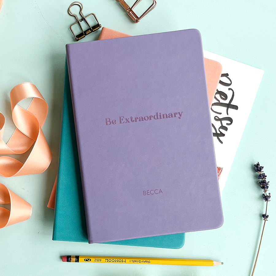 The front of the notebook features the engraved text Be Extraordinary. It is personalised with the name Becca. The lilac book is stacked on top of two other notebooks, turquoise and pale pink. Surrounding the book is peach ribbon, various stationary items and dried flowers.