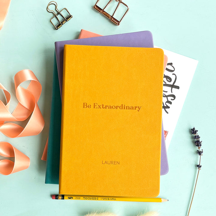 A top down image of a stack of three journals with a mustard yellow vegan leather notebook on the top resting on a pale aqua table. The front of the notebook features the words Be Extraordinary and it is personalised with the name Lauren. Surrounding the book are various stationary items and dried flowers.