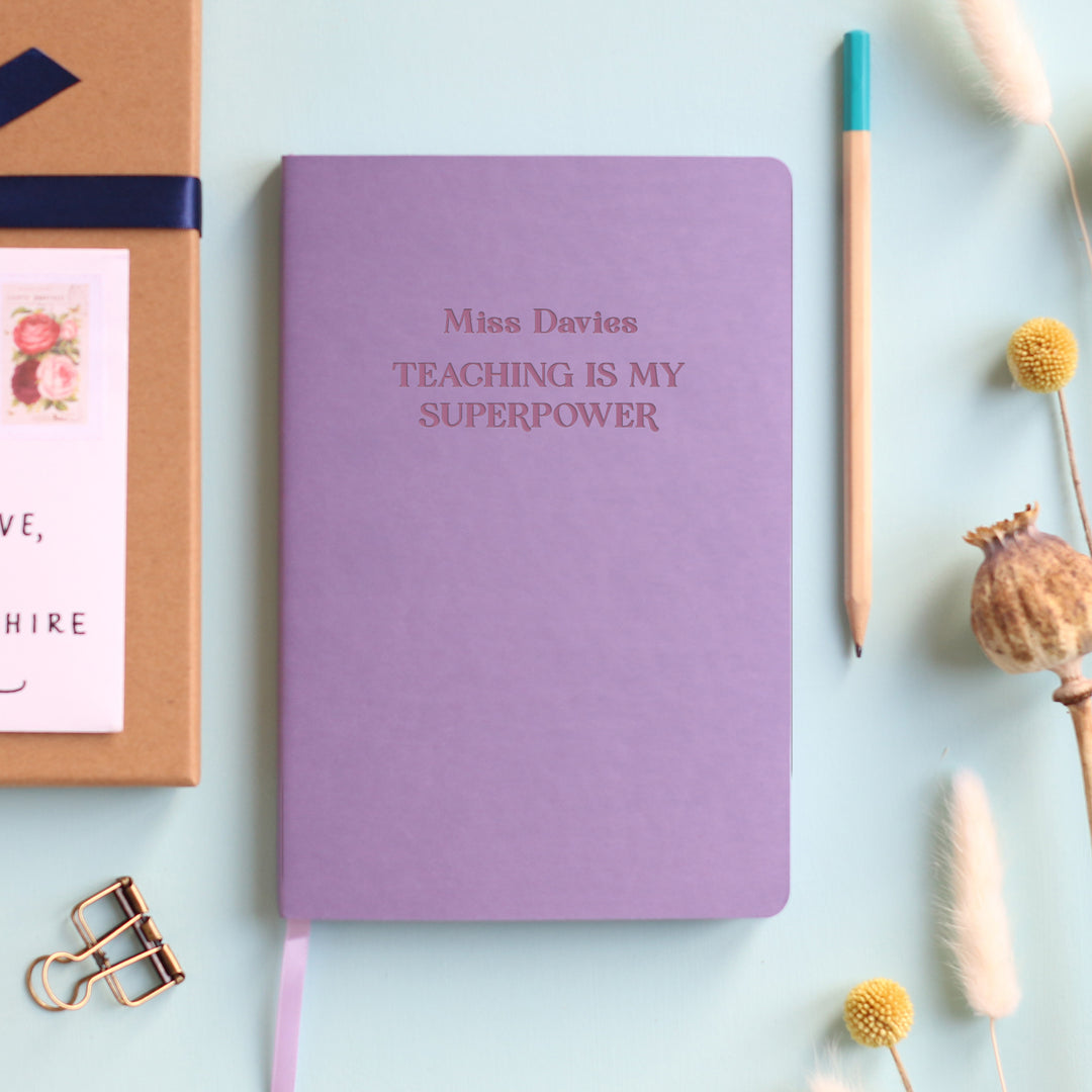 A flat lay image of a lilac vegan leather notebook resting on a pale aqua table. The front of the notebook is engraved with the phrase “Teaching is my Superpower” and is personalised with the name Miss Davies. Surrounding the book are various stationary items and dried flowers.