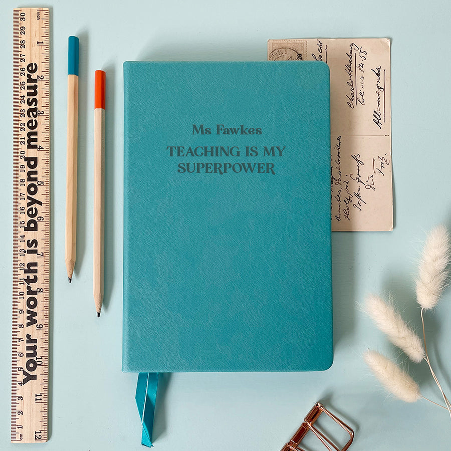 A flat lay image of a turquoise vegan leather notebook resting on a pale aqua table. The front of the notebook is engraved with the phrase “Teaching is my Superpower” and is personalised with the name Ms Fawkes. Surrounding the book are various stationery items and dried flowers.