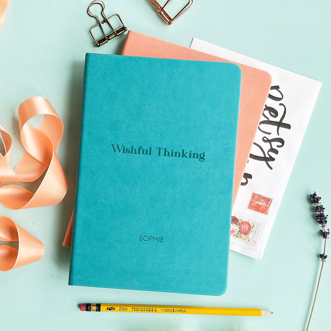 A top down image of a turquoise vegan leather notebook stacked on top of a pink notebook and resting on a pale aqua table. The front of the turquoise notebook is engraved with the phrase "Wishful Thinking" and personalised with the name Sophie. Surrounding the book are various stationary items and dried flowers.