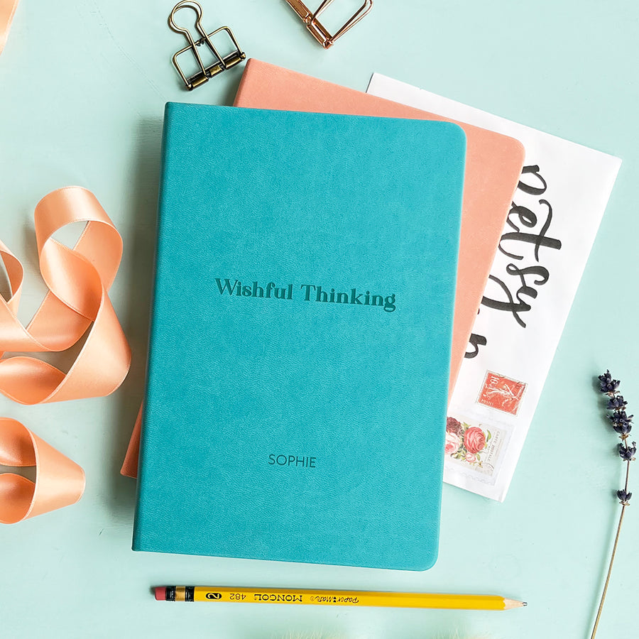 A top down image of a turquoise vegan leather notebook stacked on top of a pink notebook and resting on a pale aqua table. The front of the turquoise notebook is engraved with the phrase "Wishful Thinking" and personalised with the name Sophie. Surrounding the book are various stationary items and dried flowers.