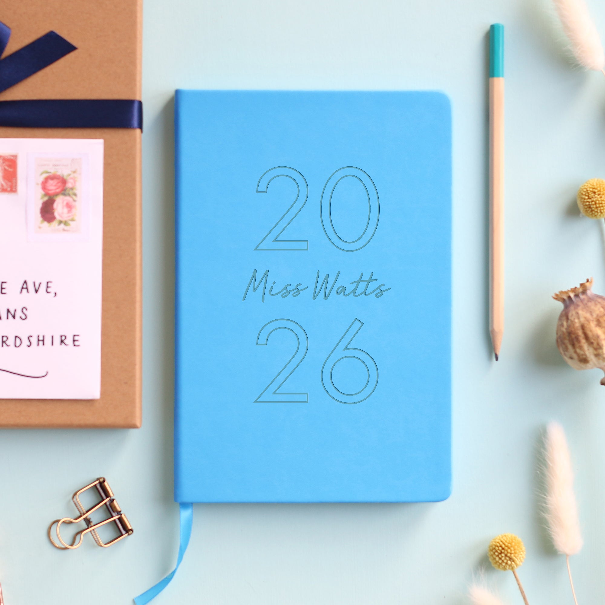 A top down image of a red vegan leather notebook resting on a pale aqua table. The front of the notebook is engraved with 2026 and the personalised text Miss Watts. Surrounding the book are various stationary items and dried flowers.