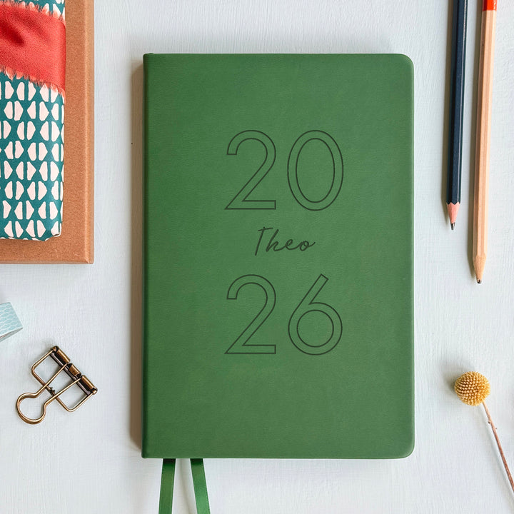 A top down image of a dark green vegan leather notebook resting on a pale aqua table. The front of the notebook is engraved with 2026 and the personalised text Theo. Surrounding the book are various stationary items and dried flowers.