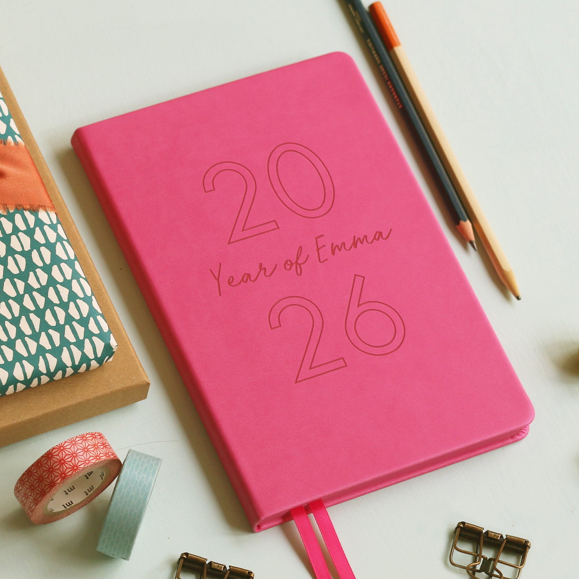A top down image of a hot pink vegan leather notebook resting on a pale aqua table. The front of the notebook is engraved with 2026 and the personalised text Year of Emma. Surrounding the book are various stationary items and dried flowers.