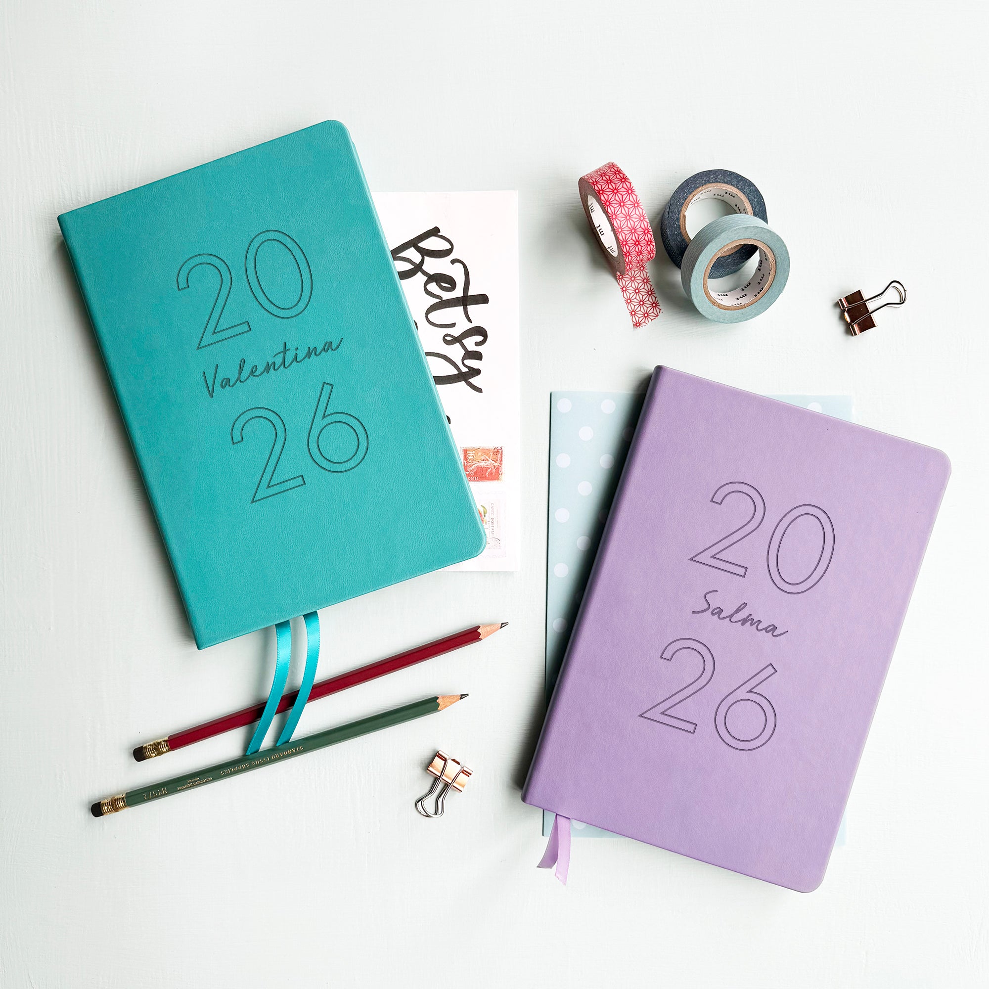 A top down image of two vegan leather notebooks, one turquoise and one lilac. They are resting on a pale aqua table. Both are engraved with 2026. The turquoise notebook is personalised the name Valentina and the Lilac book features the name Salma. Surrounding the book are various stationary items.