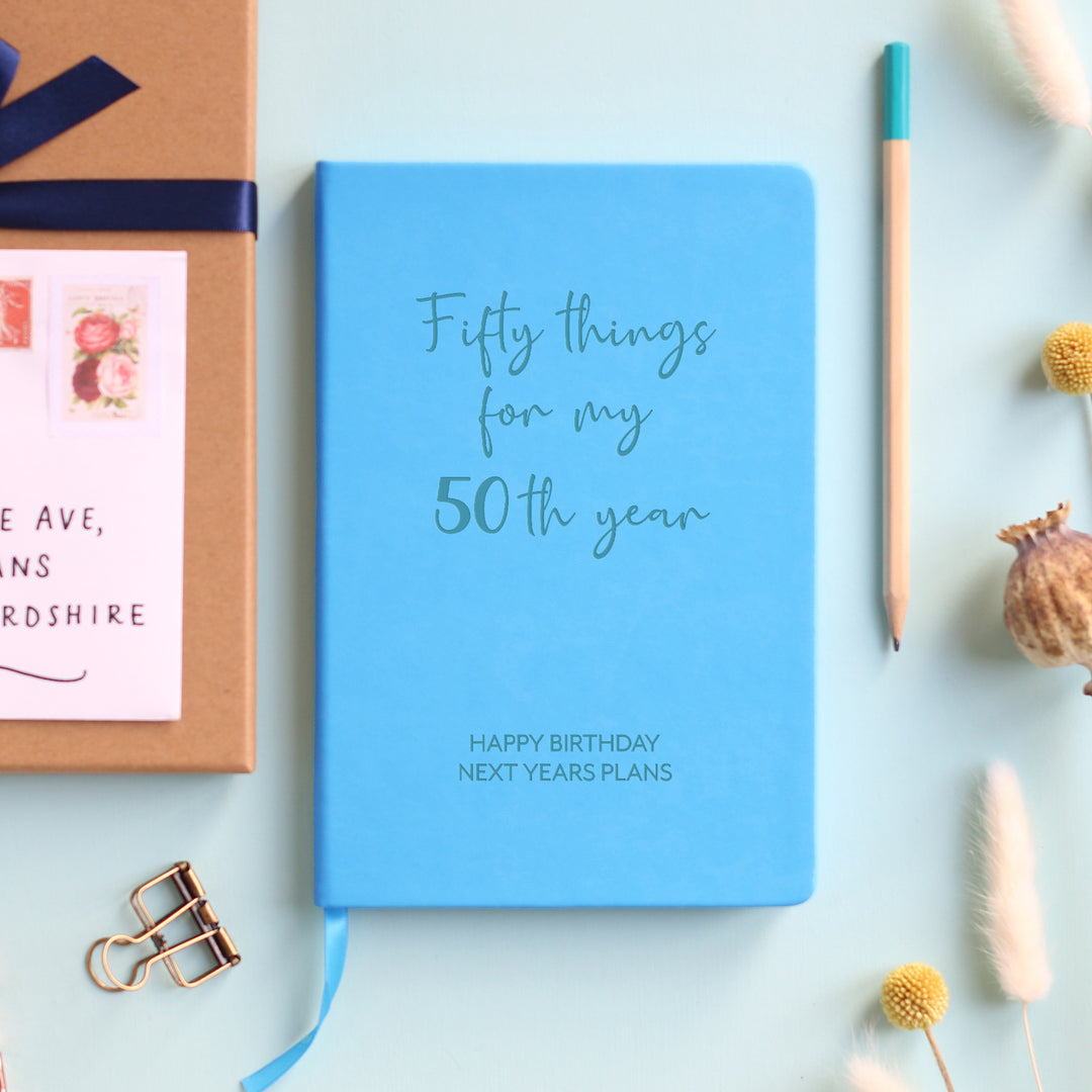 A top down image of a blue vegan leather notebook resting on a pale aqua table. The front of the notebook features the words Fifty things for my 50th year. The personalised engraving reads Happy Birthday next years plans. Surrounding the book are various stationary items and dried flowers.
