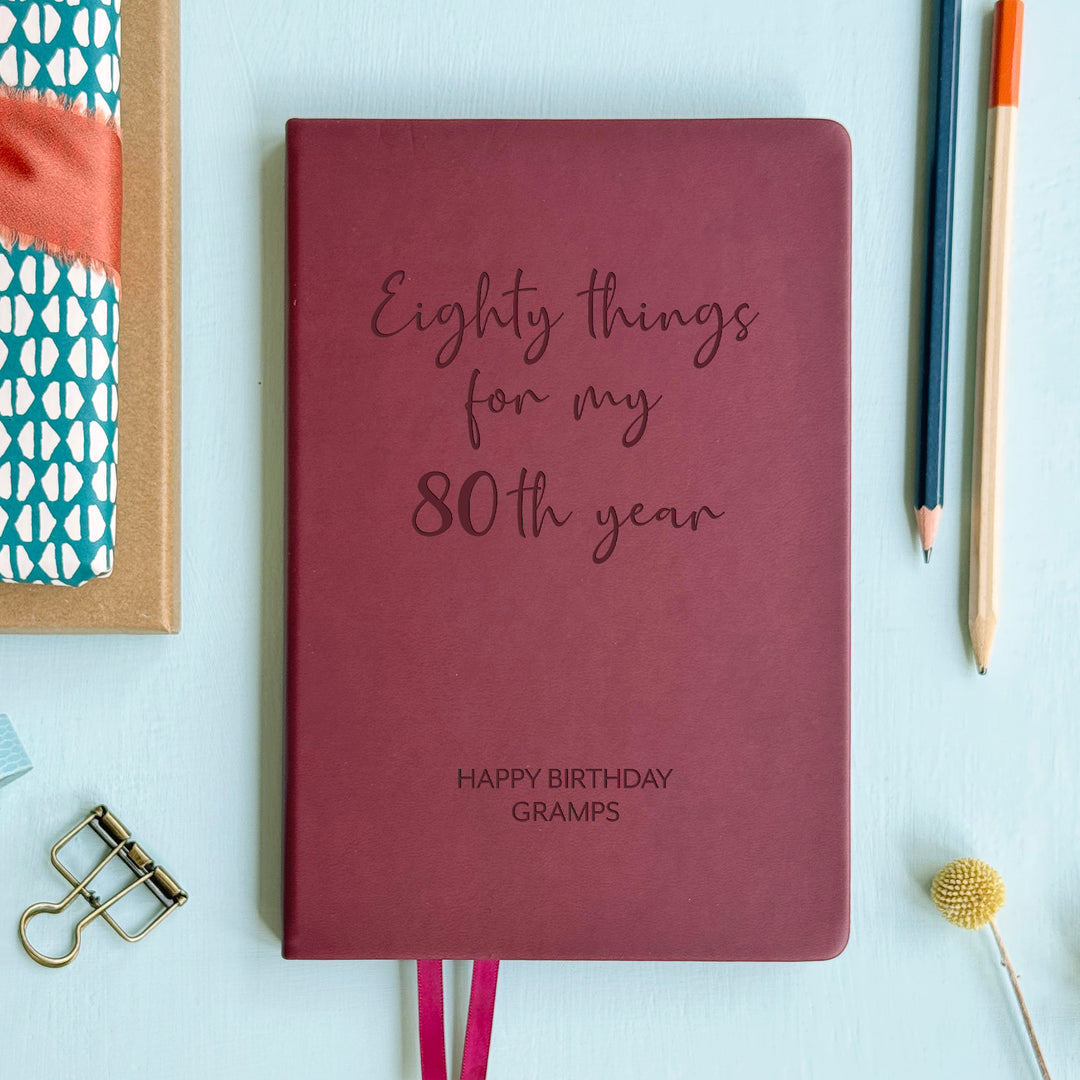 A top down image of an burgundy vegan leather notebook resting on a pale aqua table. The front of the notebook features the words eighty things for my 80th year. The personalised engraving reads Birthday Planner. Surrounding the book are various stationary items and dried flowers.
