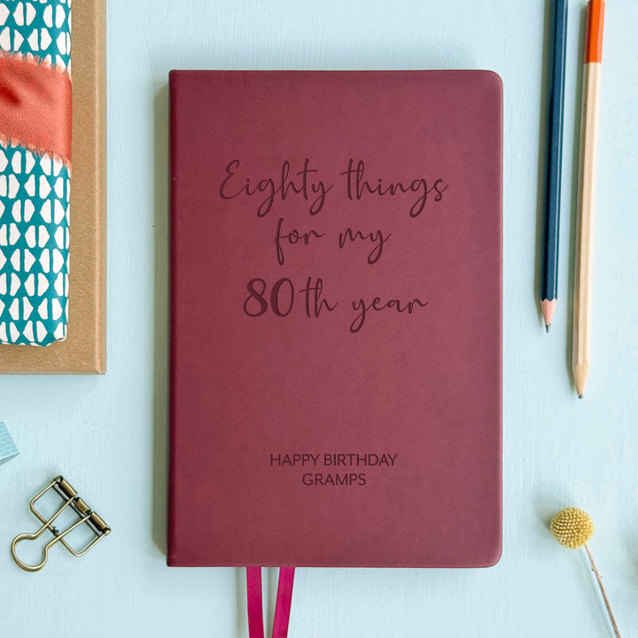A top down image of an burgundy vegan leather notebook resting on a pale aqua table. The front of the notebook features the words eighty things for my 80th year. The personalised engraving reads Birthday Planner. Surrounding the book are various stationary items and dried flowers.