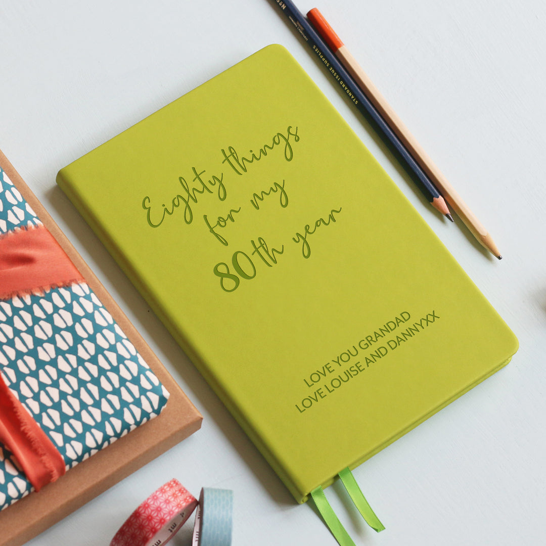 A top down image of an lime green vegan leather notebook resting on a pale aqua table. The front of the notebook features the words eighty things for my 80th year. The personalised engraving reads Birthday Planner. Surrounding the book are various stationary items and dried flowers.