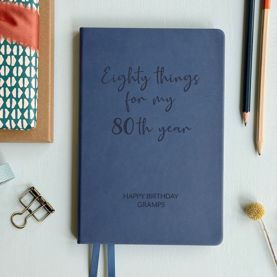 A top down image of an mid blue vegan leather notebook resting on a pale aqua table. The front of the notebook features the words eighty things for my 80th year. The personalised engraving reads Birthday Planner. Surrounding the book are various stationary items and dried flowers.