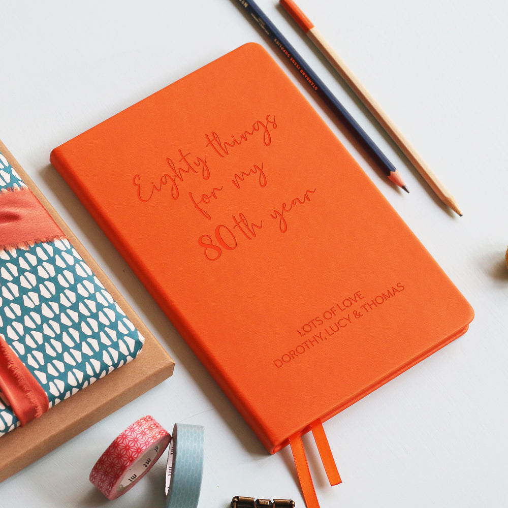 A top down image of an orange vegan leather notebook resting on a pale aqua table. The front of the notebook features the words eighty things for my 80th year. The personalised engraving reads Birthday Planner. Surrounding the book are various stationary items and dried flowers.