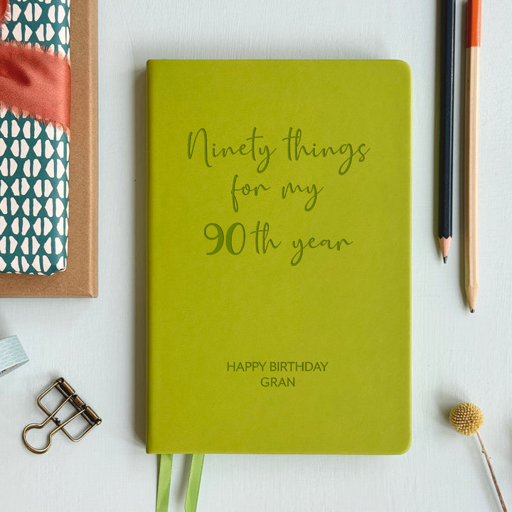 A top down image of an lime green vegan leather notebook resting on a pale aqua table. Engraved on the front of the notebook is ninety things for my 90th year. The personalised engraving reads Happy Birthday Gran.  Surrounding the book are various stationary items and dried flowers.