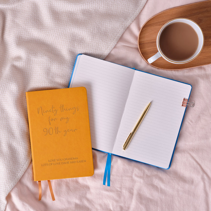 A top down image of an mustard yellow vegan leather notebook resting on a pale aqua table. Engraved on the front of the notebook is ninety things for my 90th year. The personalised engraving reads Love You Grandma Lots of Love Dave and Karen  Surrounding the book are various stationary items and dried flowers.