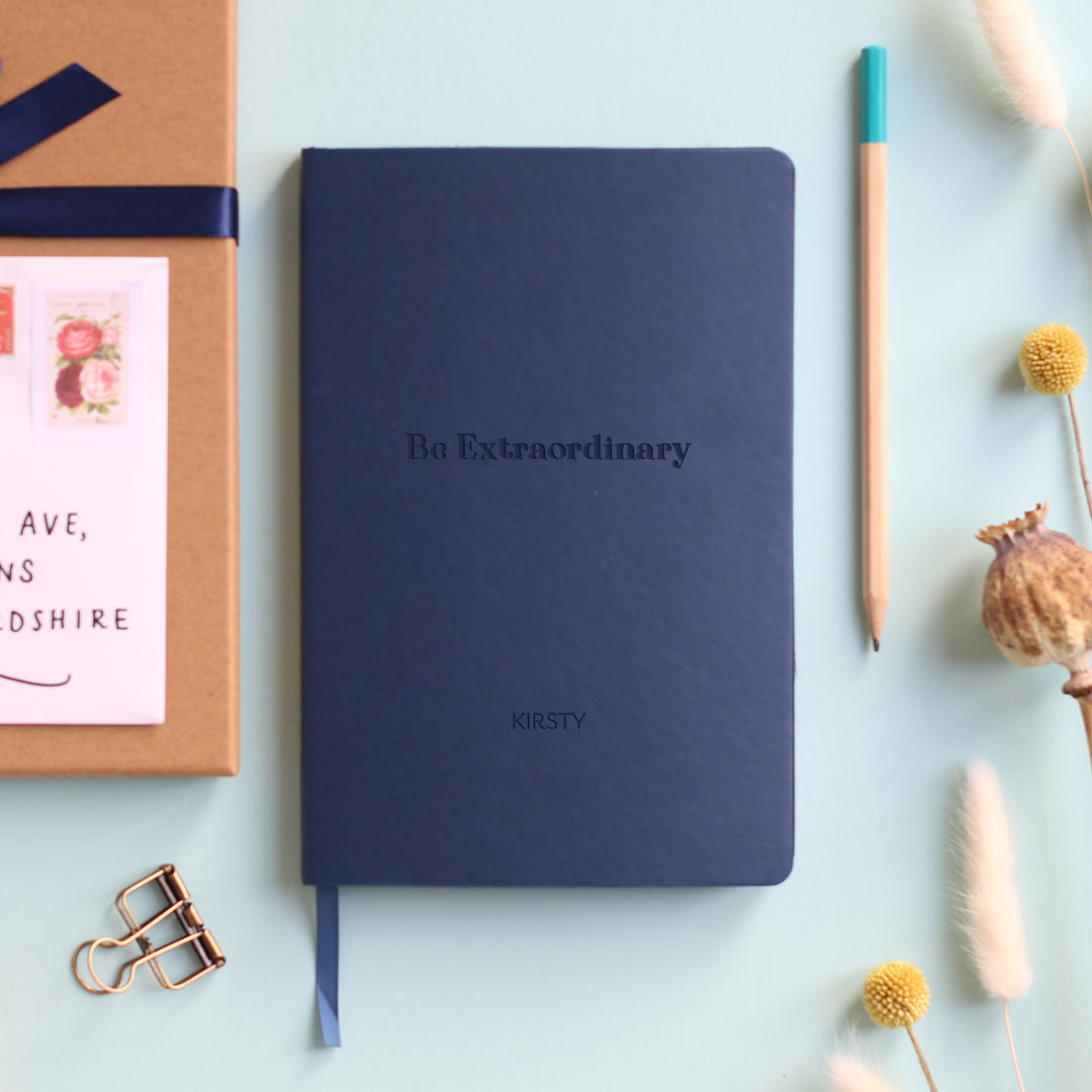 A top down image of a navy blue vegan leather notebook resting on a pale aqua table. The front of the notebook features the words Be Extraordinary. It is personalised with the name Kirsty. Surrounding the book are various stationary items and dried flower heads.